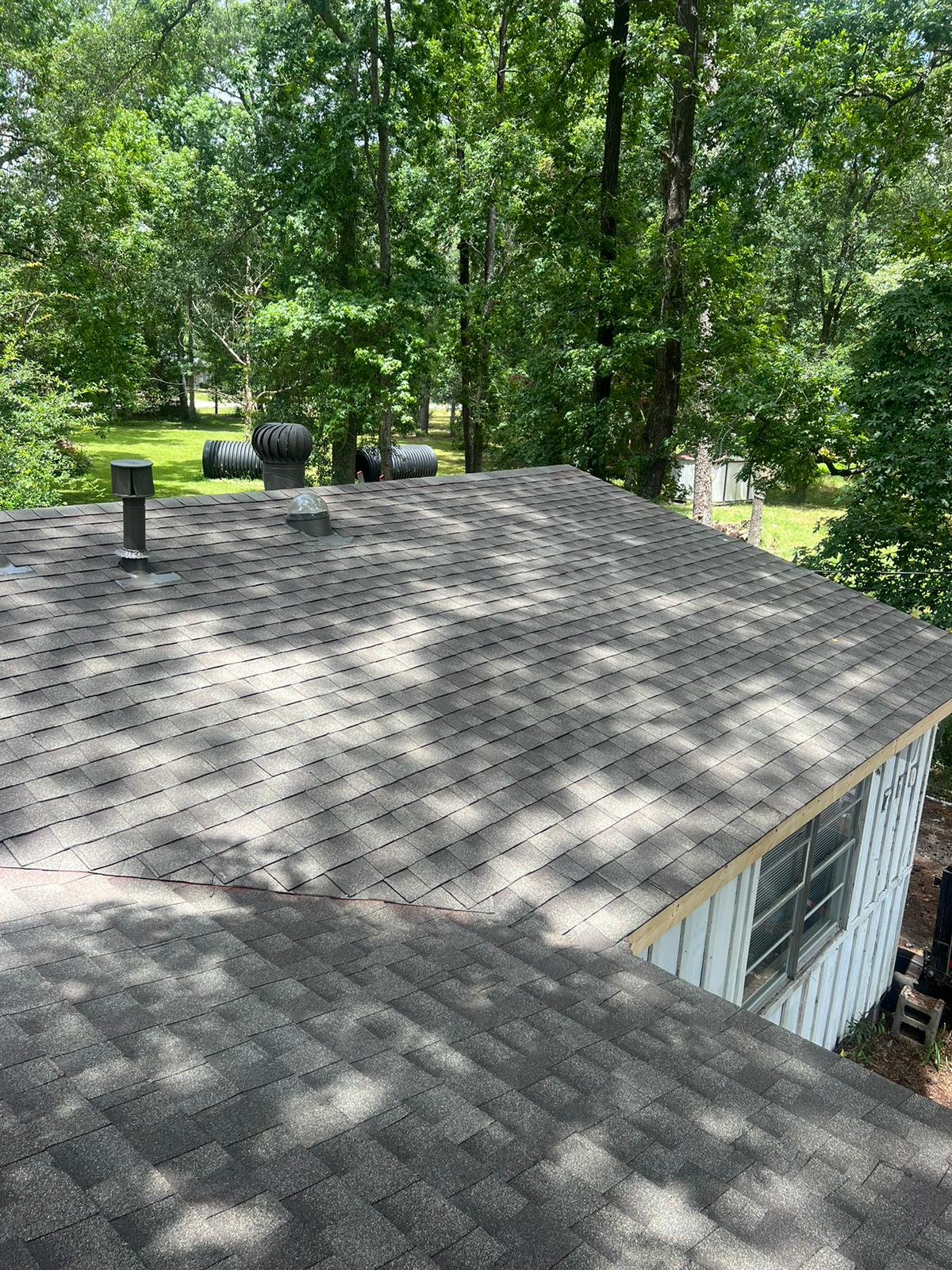 Shingled residential roof with attic vents and surrounding green trees casting shadows.