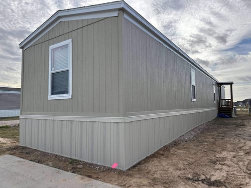 Side and end view of a beige mobile home with vertical siding, white-trimmed windows, and a small porch at the far end under a cloudy sky.