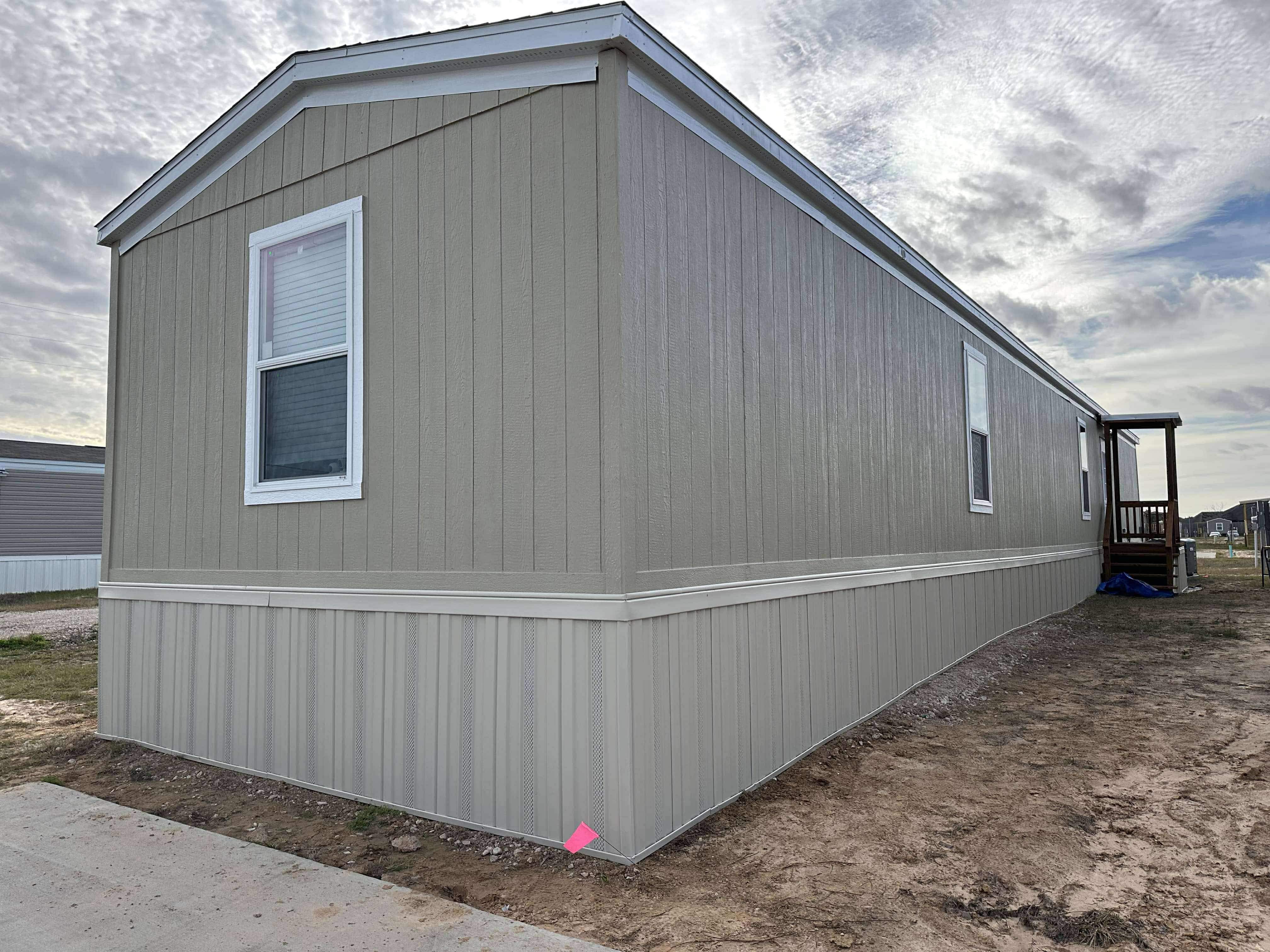 Side and end view of a beige mobile home with vertical siding, white-trimmed windows, and a small porch at the far end under a cloudy sky.