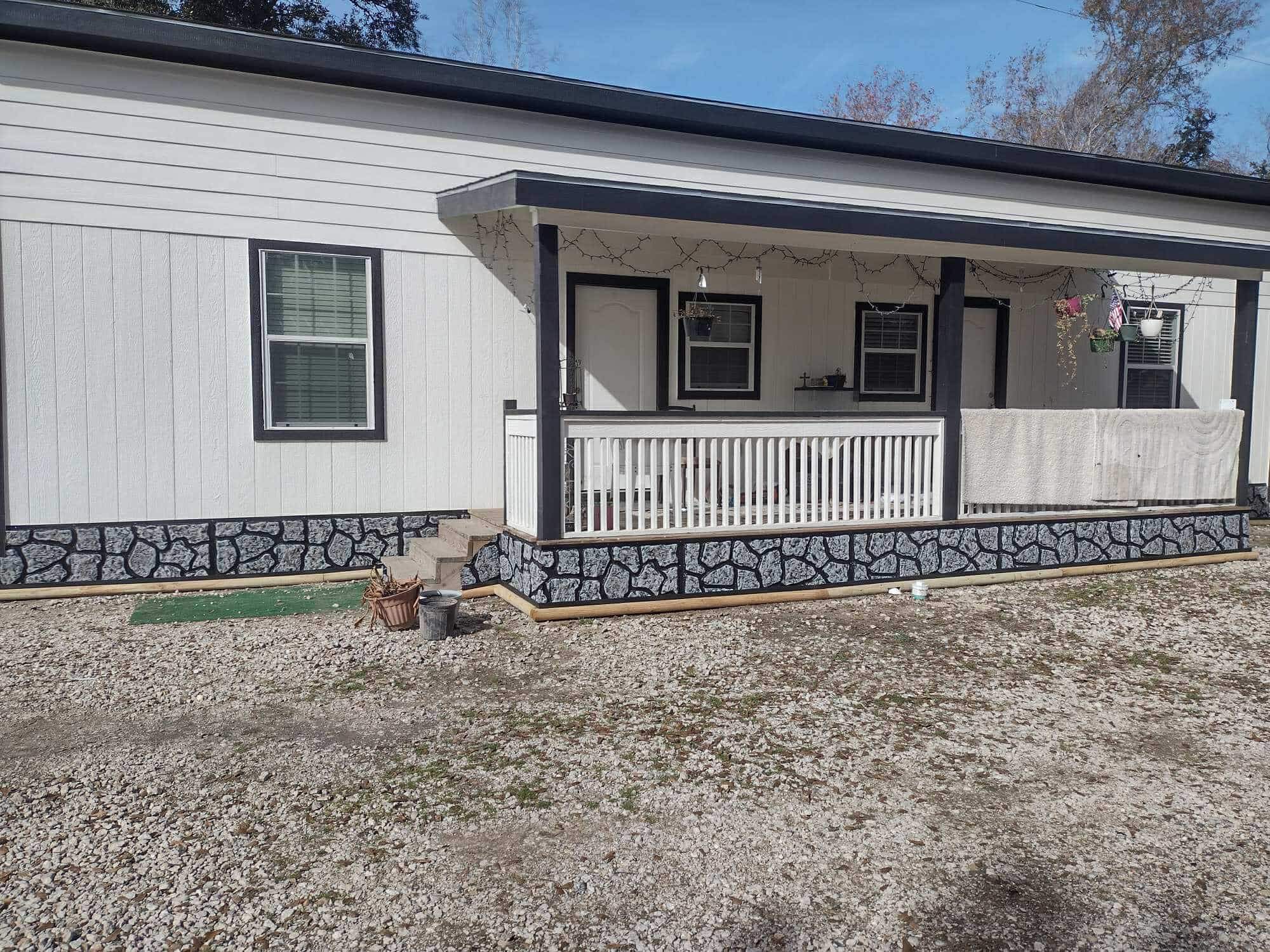 White house with black trim siding, a covered porch with railings, hanging plants, and stone patterned skirting, facing a gravel driveway.