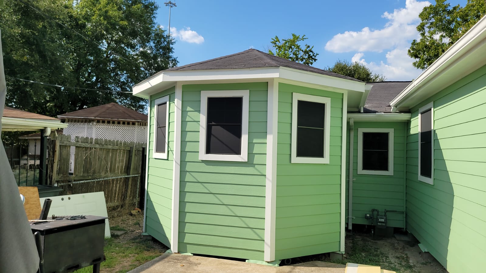 Small green octagonal exterior shed or room attached to a house with black windows and white trim under a gray roof on a sunny day.