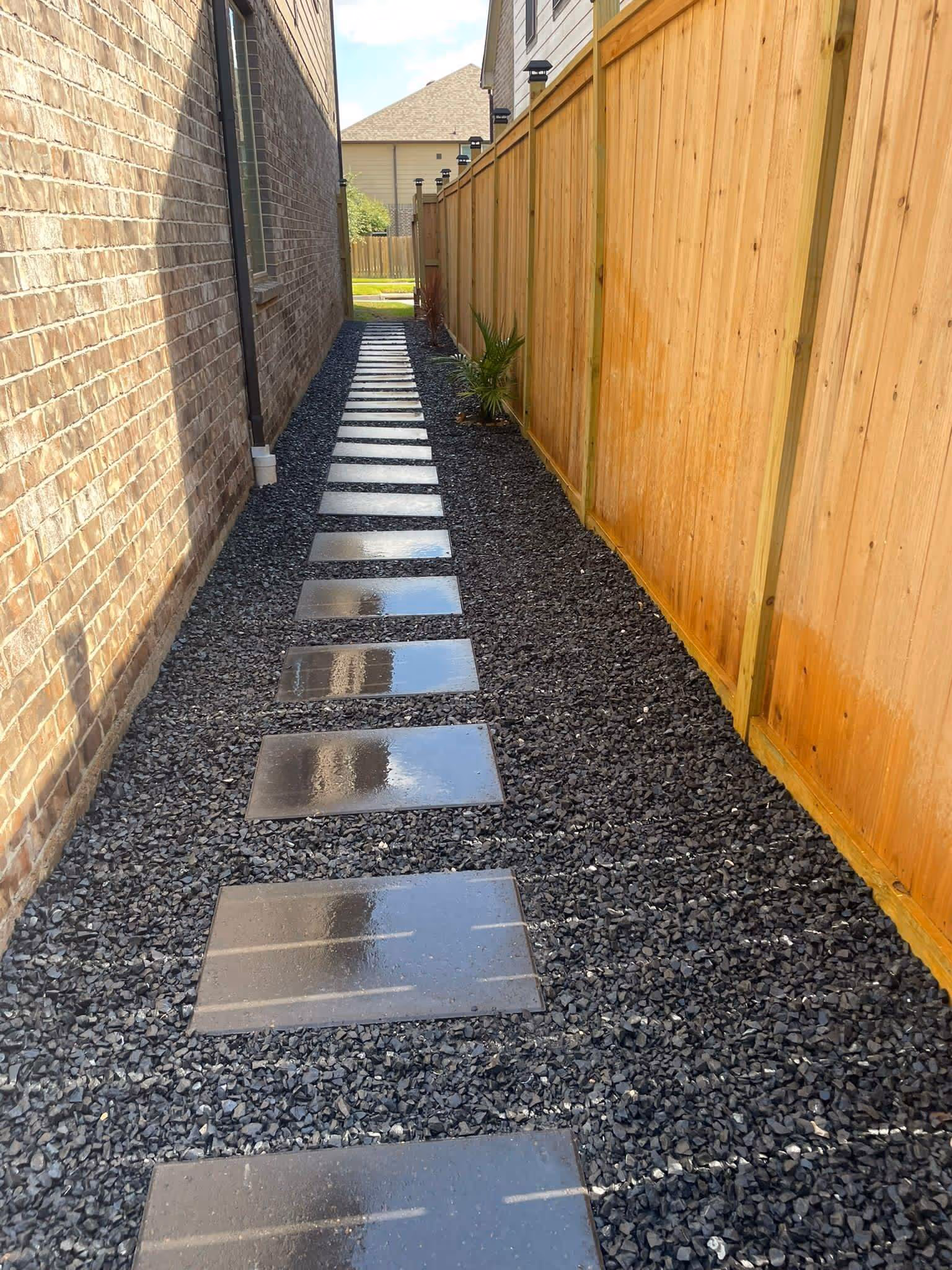 Narrow outdoor pathway with wet rectangular stepping stones set in black gravel between a brick wall and wooden fence, with small plants near the fence.