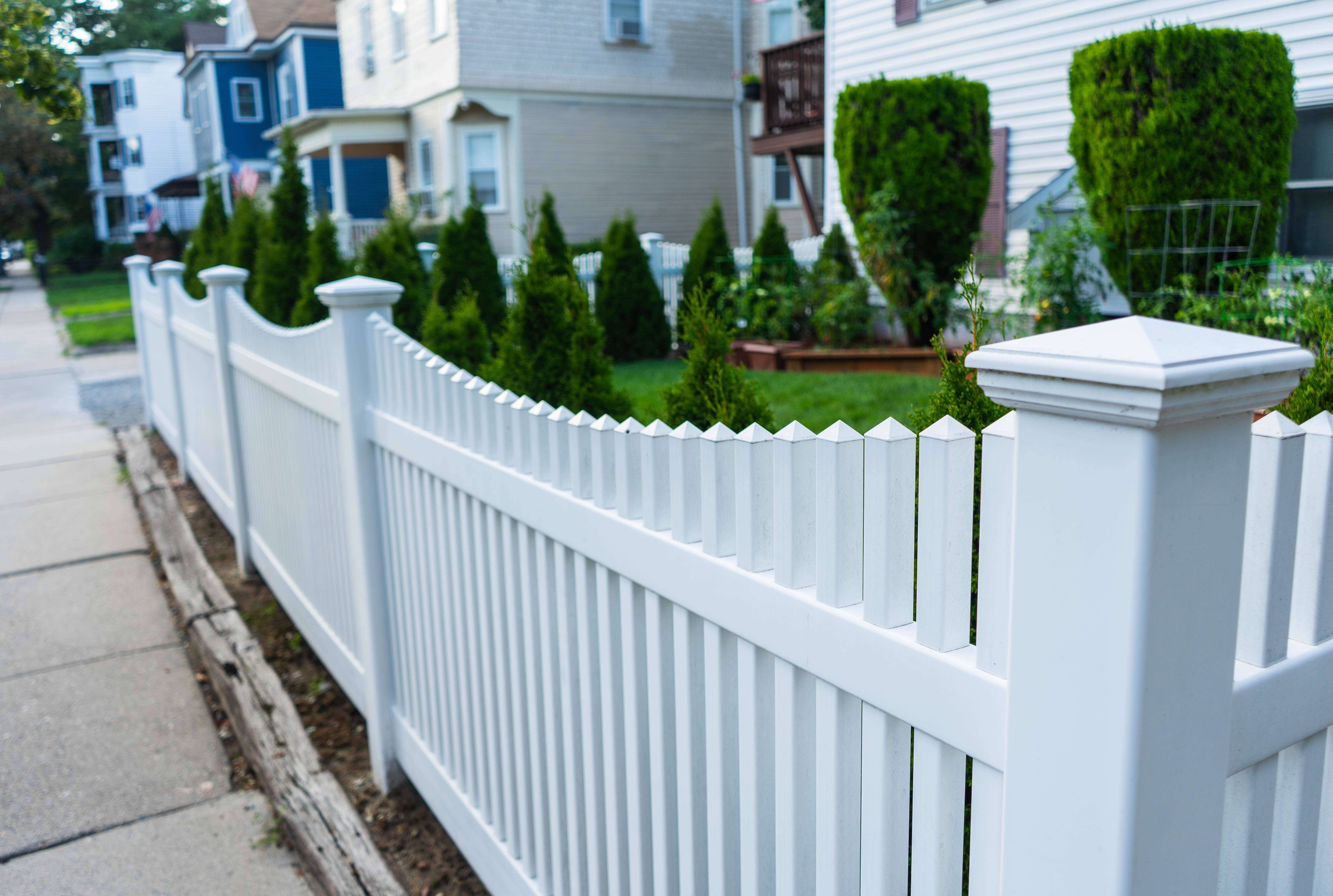 Long white picket fence bordering a sidewalk with green shrubs and residential houses in the background.