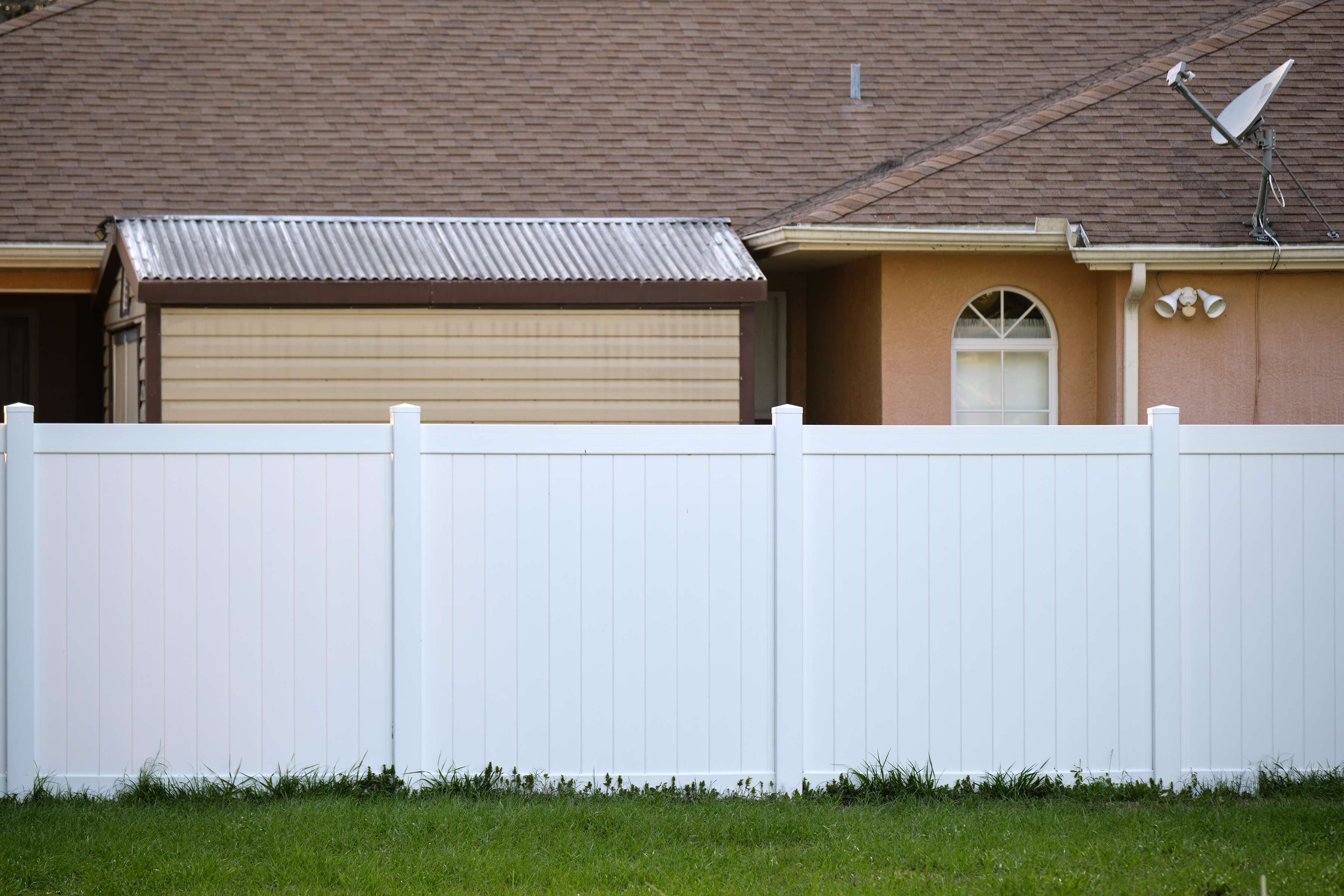 White vinyl privacy fence in front of a tan house and a shed with a corrugated metal roof.